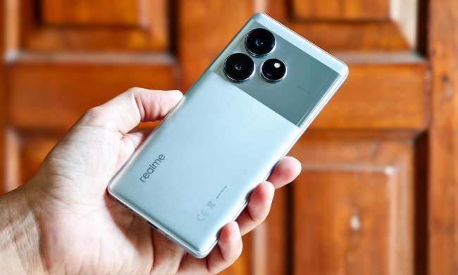 Realme GT 6 Android phone in silver color held in hand in front of a wooden door.